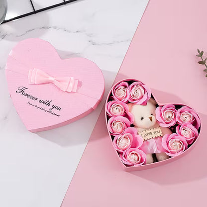 Heart-shaped pink gift box with a teddy bear and flowers on a white marble surface.