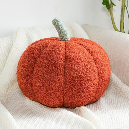 Fuzzy orange pumpkin pillow on a white blanket with a plant in the background