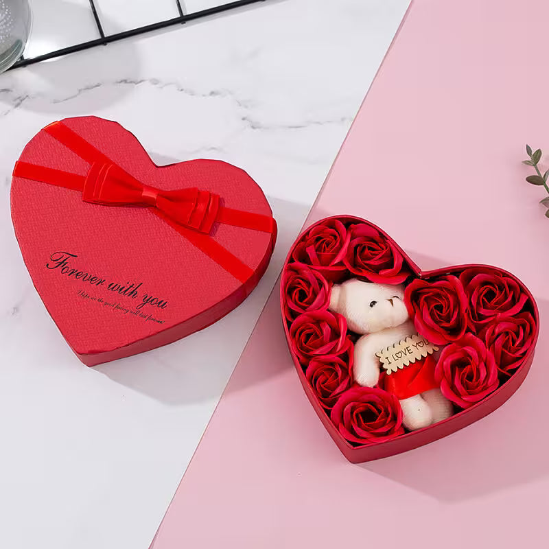 Heart-shaped gift box with a teddy bear and red roses on a pink surface.