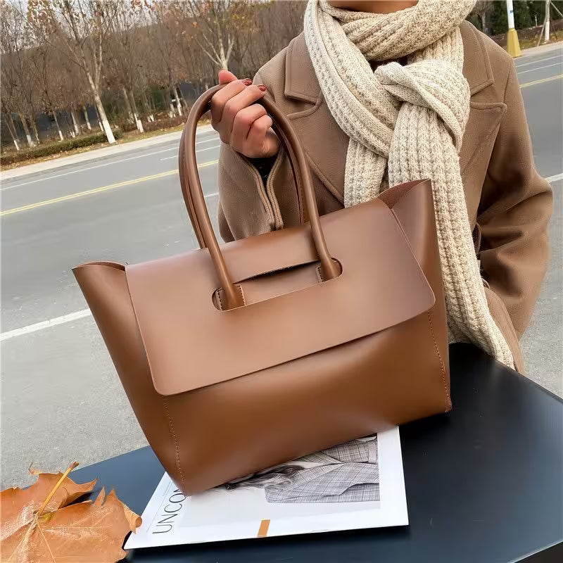 Brown handbag held by a person wearing a beige coat and scarf, with a magazine underneath on a road.
