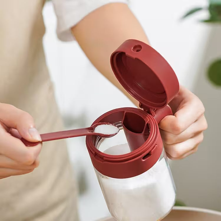 Person using a red measuring spoon to add a white substance into a clear container with a red lid.