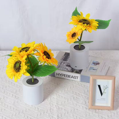 Two small sunflower arrangements in white pots on a textured surface with a magazine and photo frame in the background.