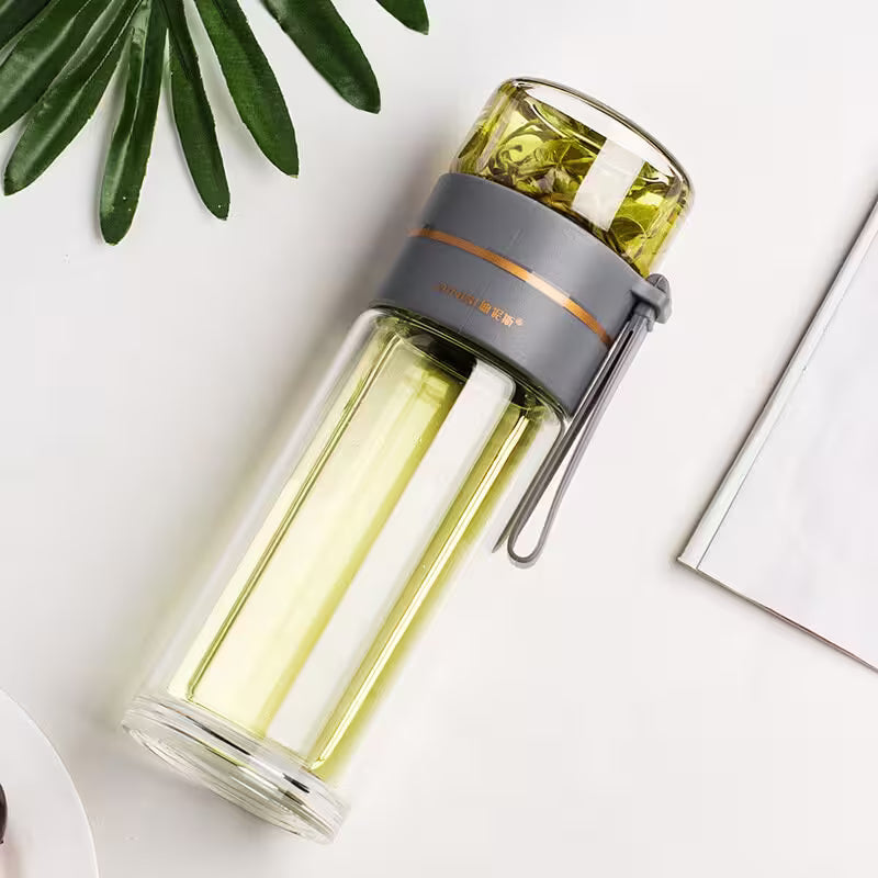 Clear glass water bottle with gray lid on a white surface with green leaves