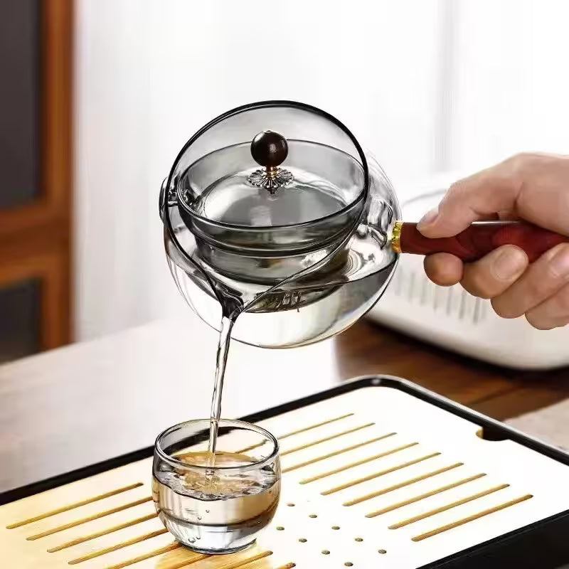 Person pouring liquid from a glass container into a small glass on a wooden surface.