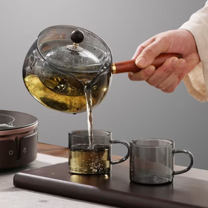 Tea being poured from a glass teapot into two cups on a wooden surface.