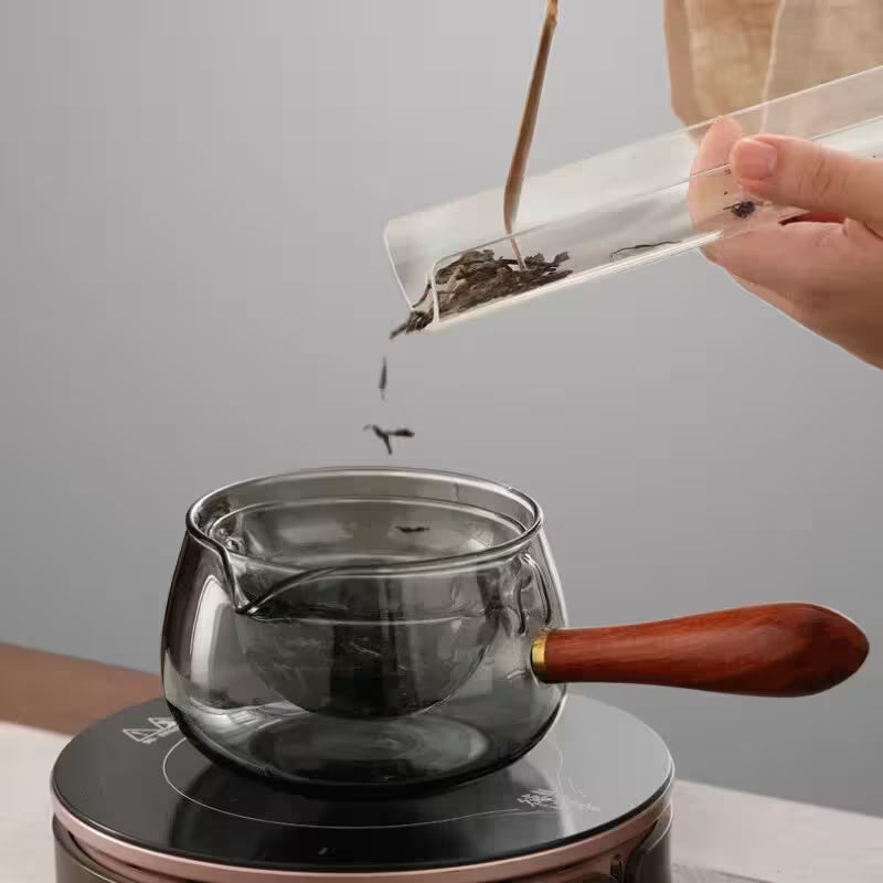 Person pouring tea leaves into a glass teapot with a wooden handle on a gray background