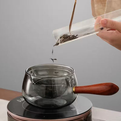 Person pouring tea leaves into a glass teapot with a wooden handle on a gray background
