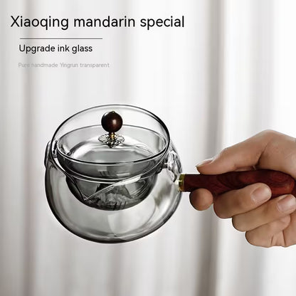 Clear glass teapot with wooden handle held by a hand against a white background