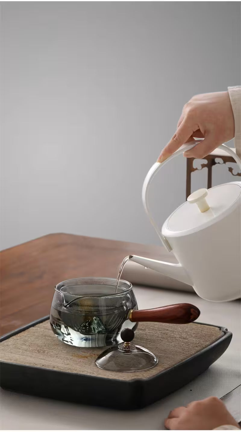 Person pouring tea from a white teapot into a glass cup on a wooden tray.