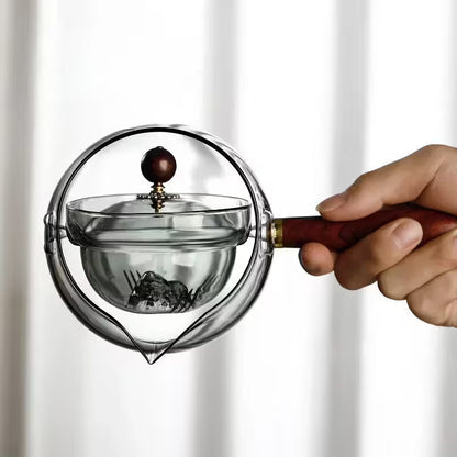 Hand holding a clear glass tea infuser with a wooden handle against a white background