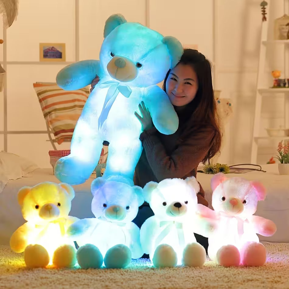 Woman holding a large glowing blue teddy bear with other colorful teddy bears on a carpeted floor.