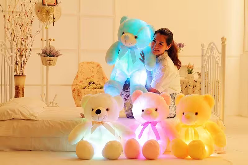 Four LED teddy bears in different colors sitting on a bed with a person in the background.