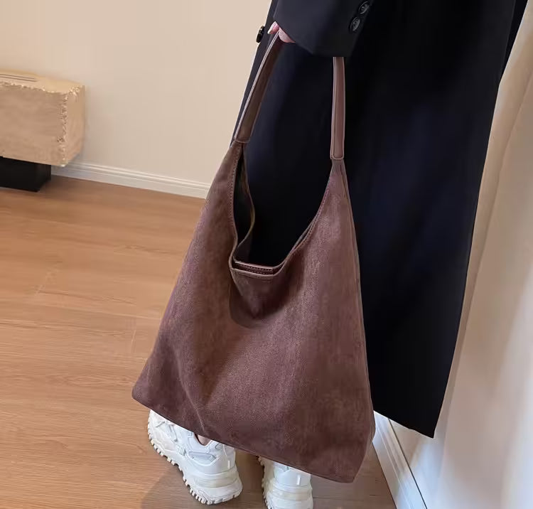 Brown suede handbag held by a person wearing a black coat and white shoes on a wooden floor.