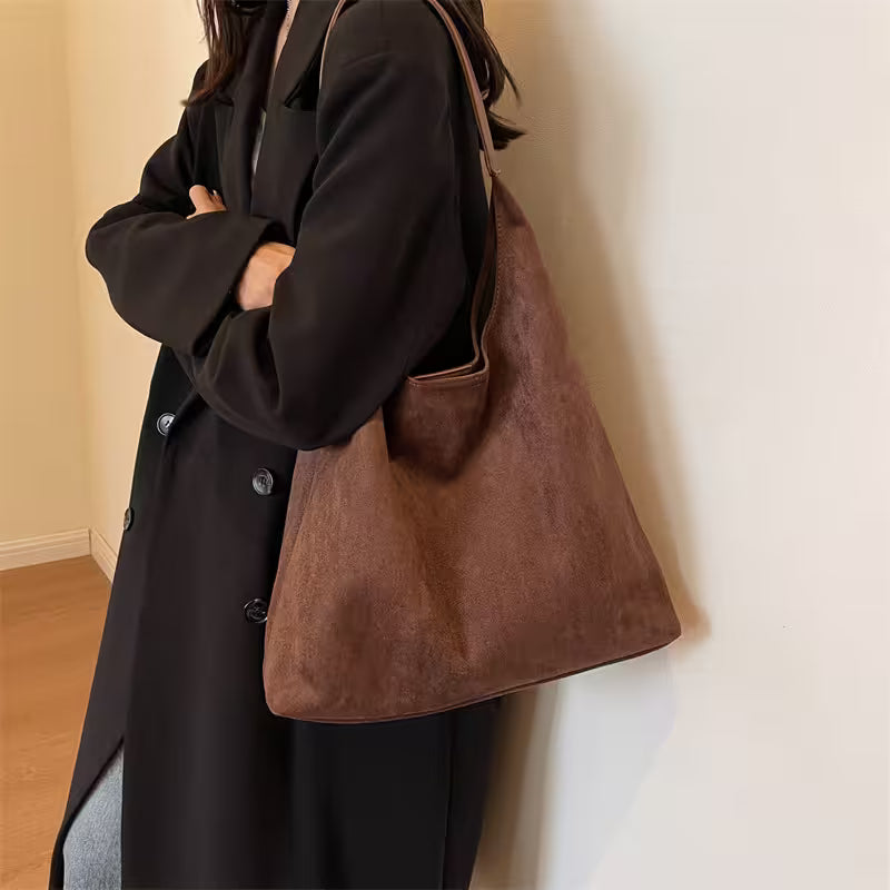 Person wearing a black coat holding a brown leather bag against a plain wall.