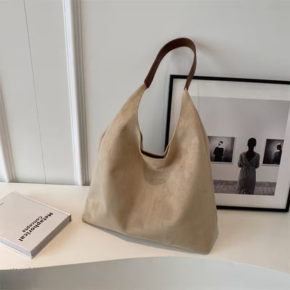 Beige handbag on a white surface with a book and framed pictures in the background