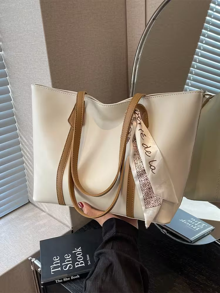 Beige handbag with brown handles on a surface with books and a scarf.