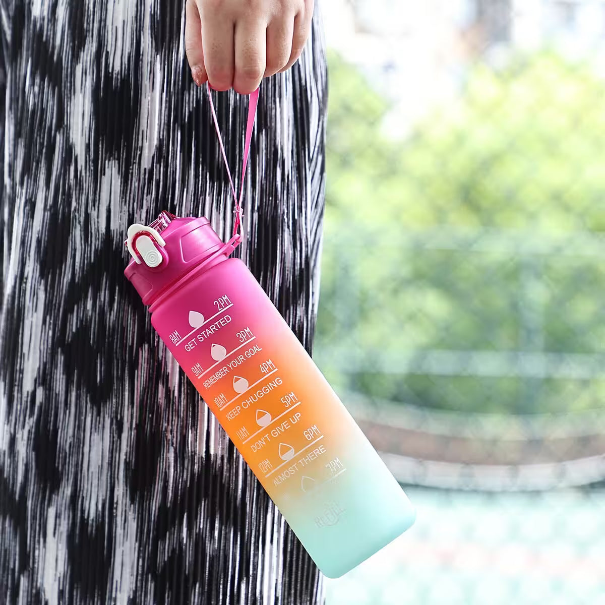 Gradient pink to blue water bottle with measurement markings held by a person outdoors.