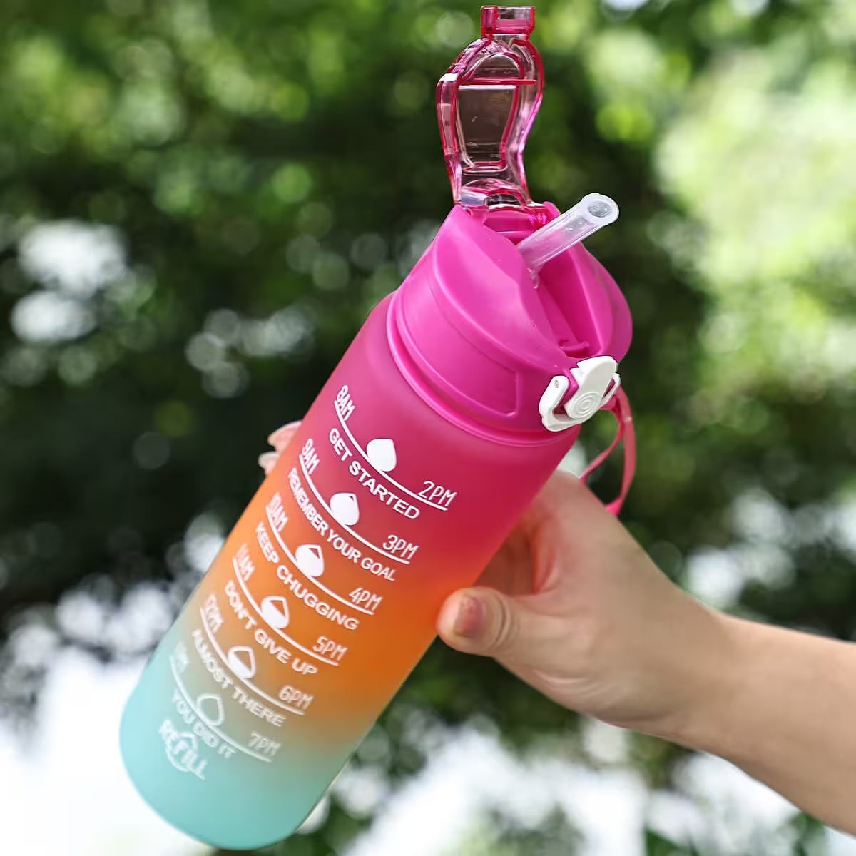 Gradient pink to blue water bottle with measurement markings held by a hand against a blurred green background