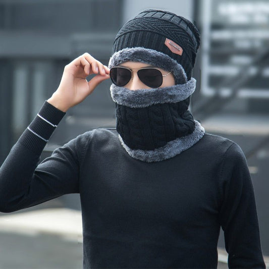 Person wearing a black knit hat and scarf with sunglasses, standing against a blurred background.