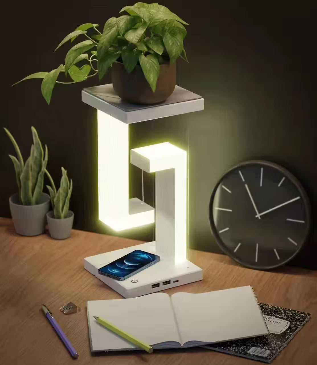 Modern desk lamp with phone charging station on a wooden surface with plants and clock.